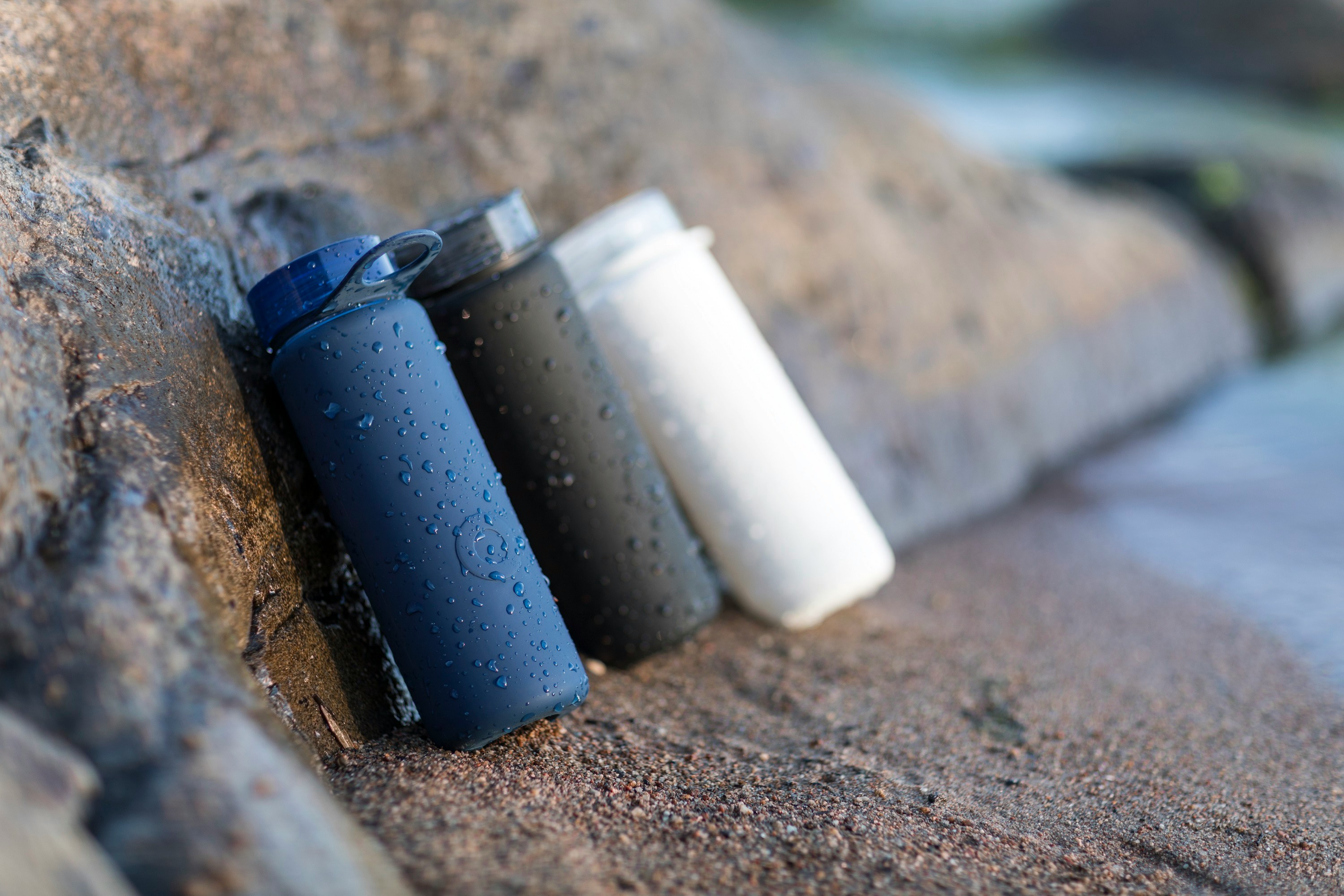 The image used in Aydya Limited customer story. It shows three water bottles in different colors (blue, black, and white) resting on rocks near a water body, with droplets of water on them.